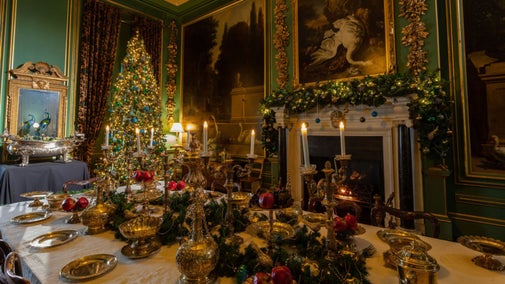 Image showing Christmas decorations inside the Hondecoeter dining room at Belton, with large tree in the corner dressed in blue and green baubles. The table has a silver gilt collection with a decorated green garland, with one to match on the mantlepiece.